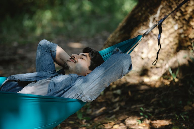 Young bearded male with closed eyes resting in hammock leaning on hands in forest