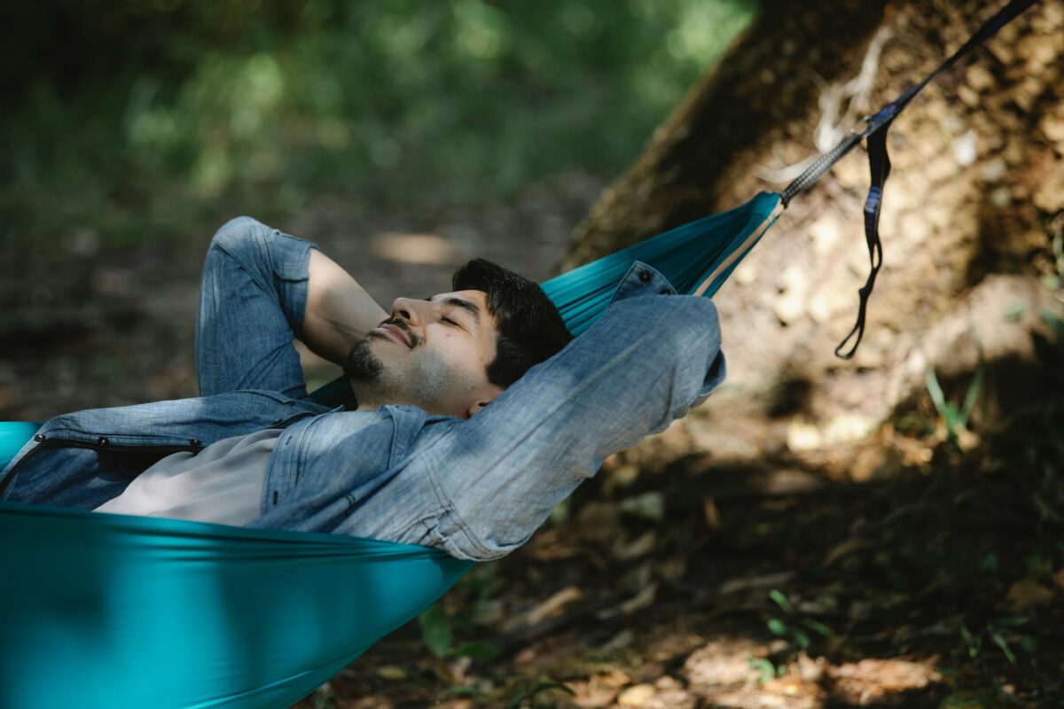 Young bearded male with closed eyes resting in hammock leaning on hands in forest