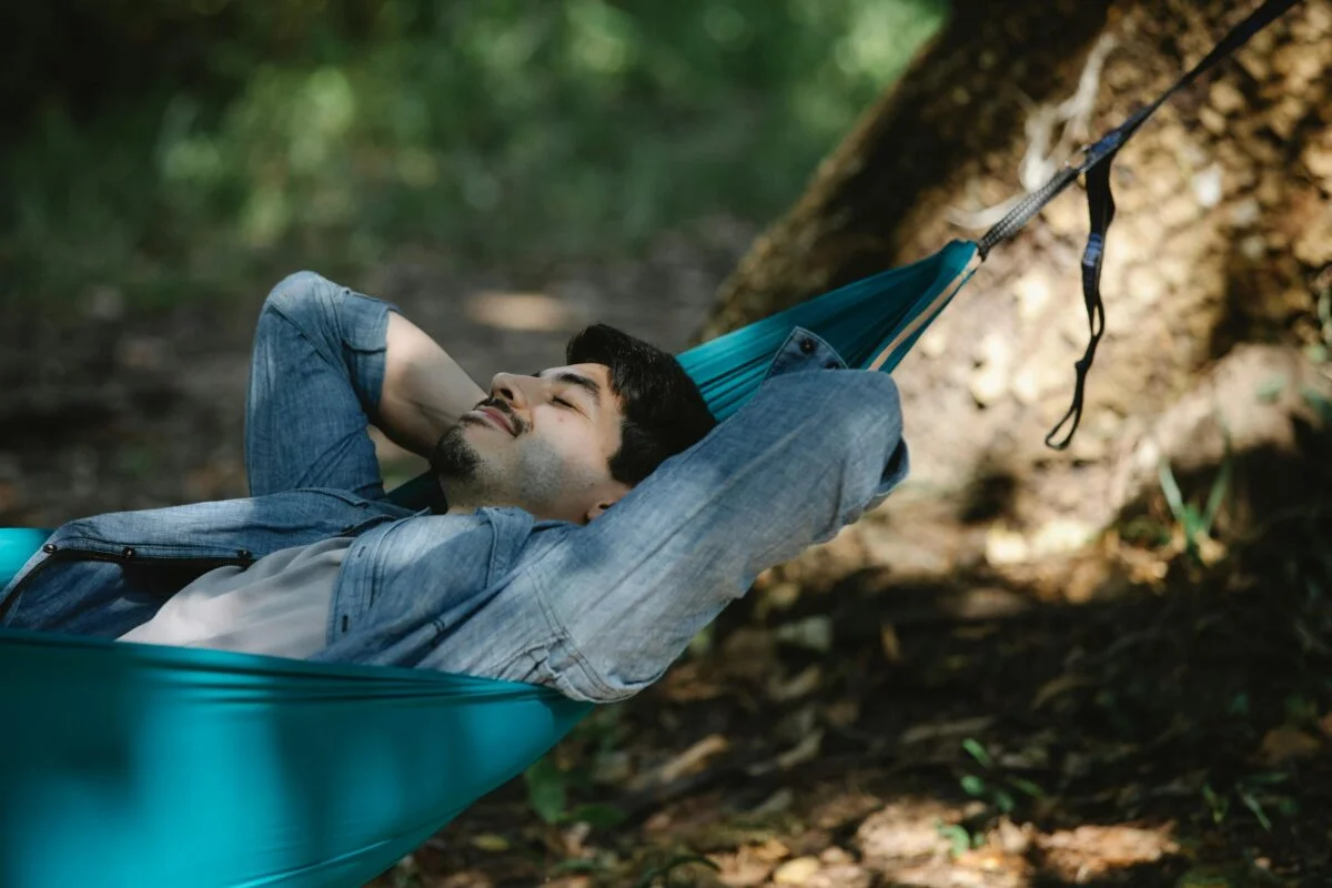 Young bearded male with closed eyes resting in hammock leaning on hands in forest